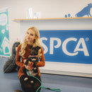 A woman kneels on a blue carpeted floor in front of a large blue and white SPCA sign, holding a small black and white dog. A green leash lies on the floor nearby, with a phone beside her.