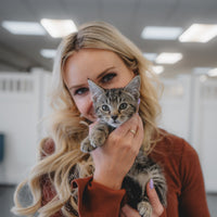 A woman with long blonde hair holds a small tabby kitten close to her face. She is wearing a rust-brown long-sleeve top, and the background shows a softly blurred indoor space with ceiling lights.