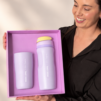 A woman holds a lavender gift box containing a matching insulated bottle and tumbler set from the brand MOOSE COPOAL. The set features a light purple tumbler and bottle with a lavender lid and a yellow accent on the bottle cap, set against a warm beige background with soft leaf shadows.